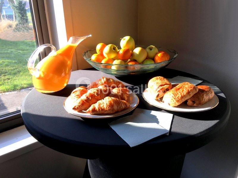 Assortiment de viennoiseries, carafe de jus d'orange et corbeille de fruits sur une table près d'une fenêtre ensoleillée.