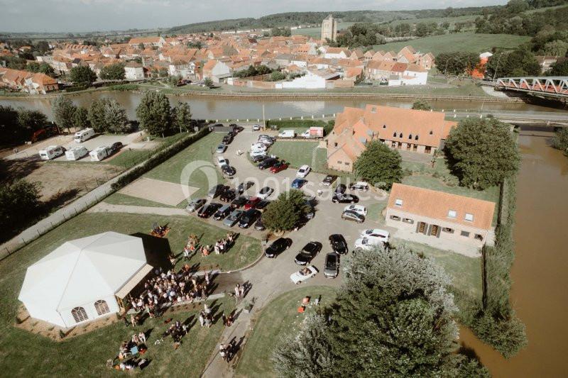 Vue aérienne d'un événement en plein air avec des bâtiments en briques, des voitures garées et un paysage rural environnant.