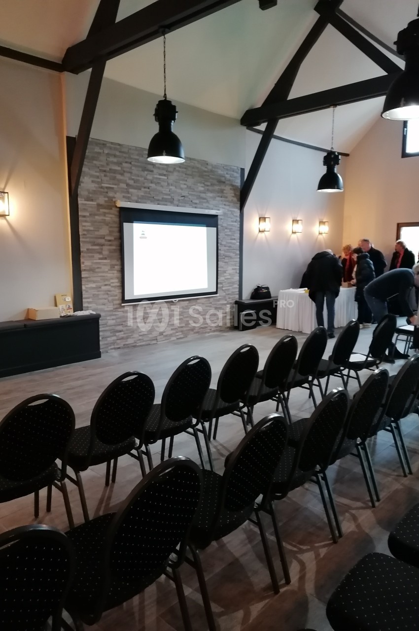Salle de conférence avec des rangées de chaises noires, un écran de projection allumé et quelques personnes debout.