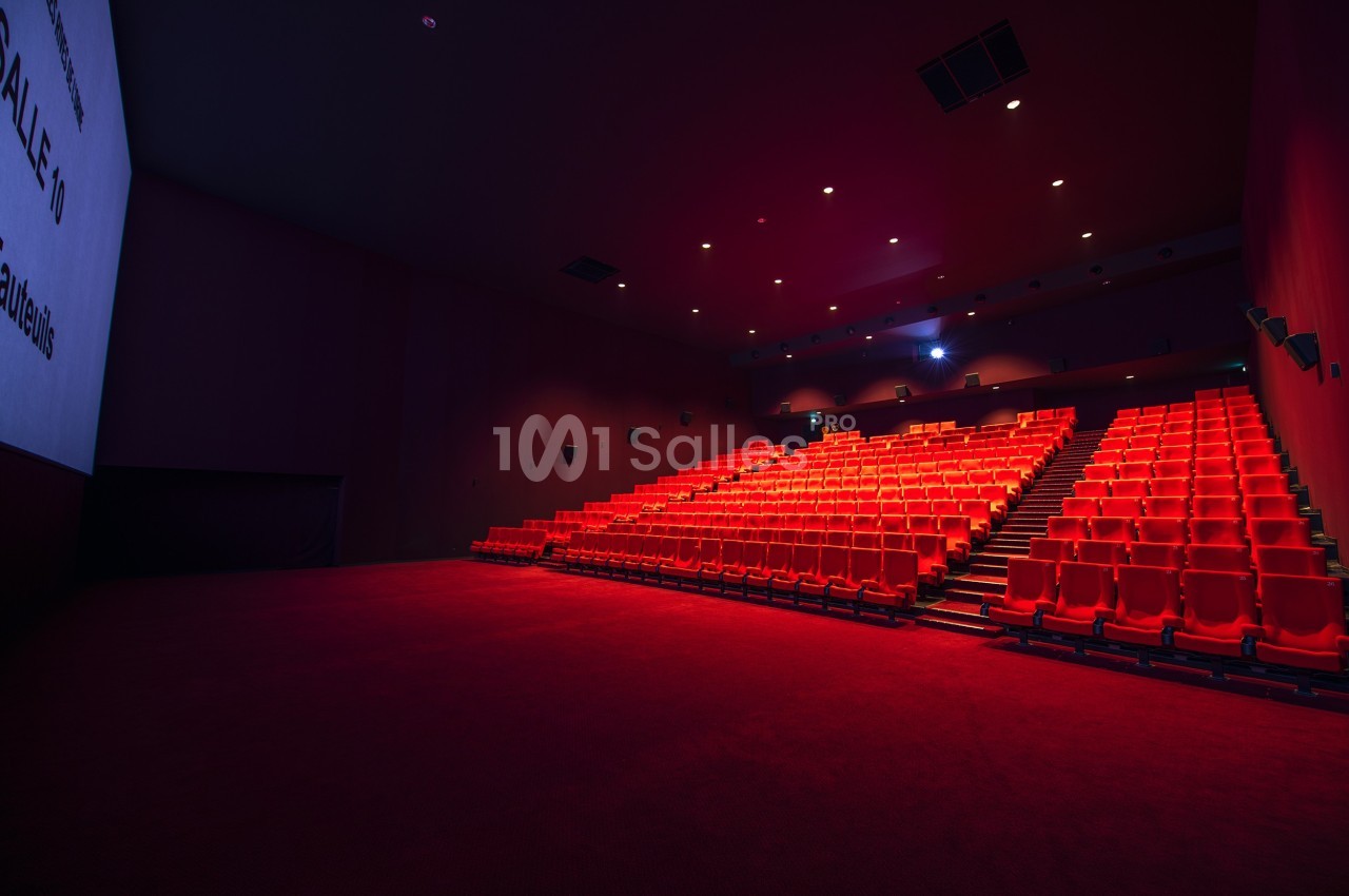 Salle de cinéma vide avec rangées de sièges rouges et écran blanc sur la gauche.