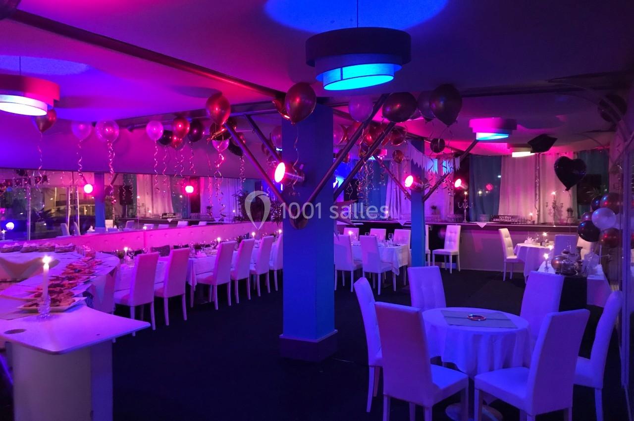 Salle décorée pour un événement avec des ballons, des tables dressées et un éclairage coloré rose et bleu.