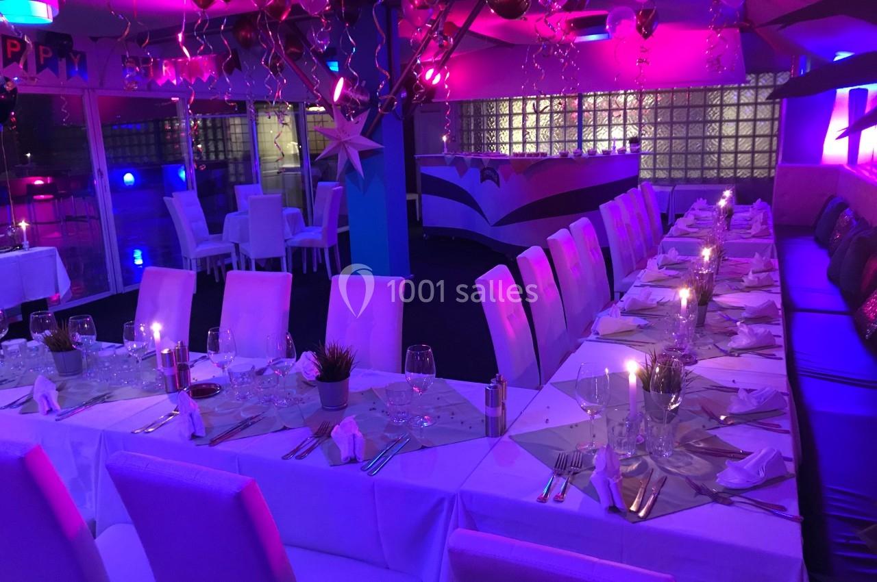 Salle décorée pour un événement, avec tables dressées, chaises blanches, ballons suspendus et éclairage coloré.