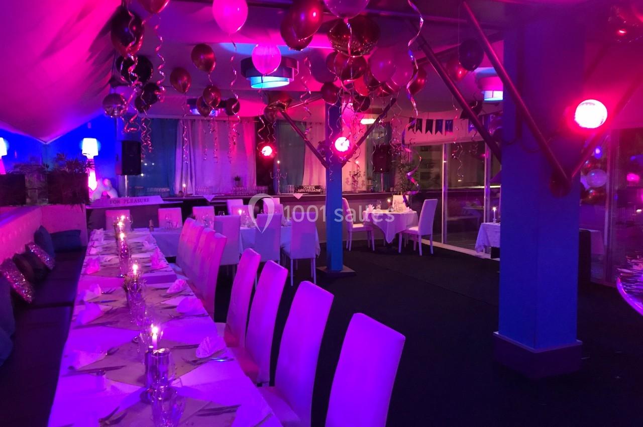 Salle décorée pour un événement festif avec tables dressées, ballons suspendus et éclairage coloré.