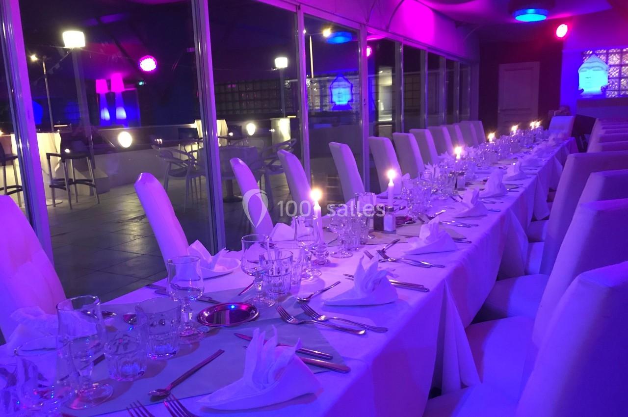 Table dressée avec nappes blanches, couverts et verres, dans une salle éclairée par des lumières violettes et bleues.