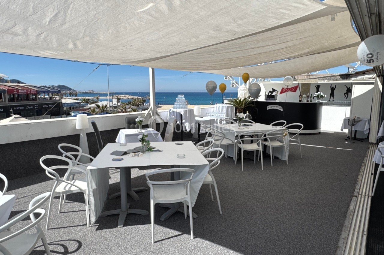 Terrasse ensoleillée avec tables et chaises blanches, vue sur la mer, décorations et ombrage par des voiles tendues.
