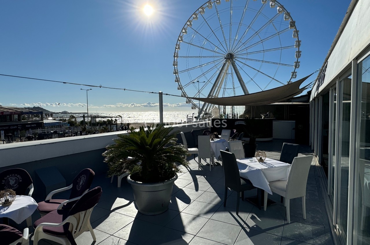 Terrasse ensoleillée avec tables dressées, vue sur une grande roue et la mer en arrière-plan.