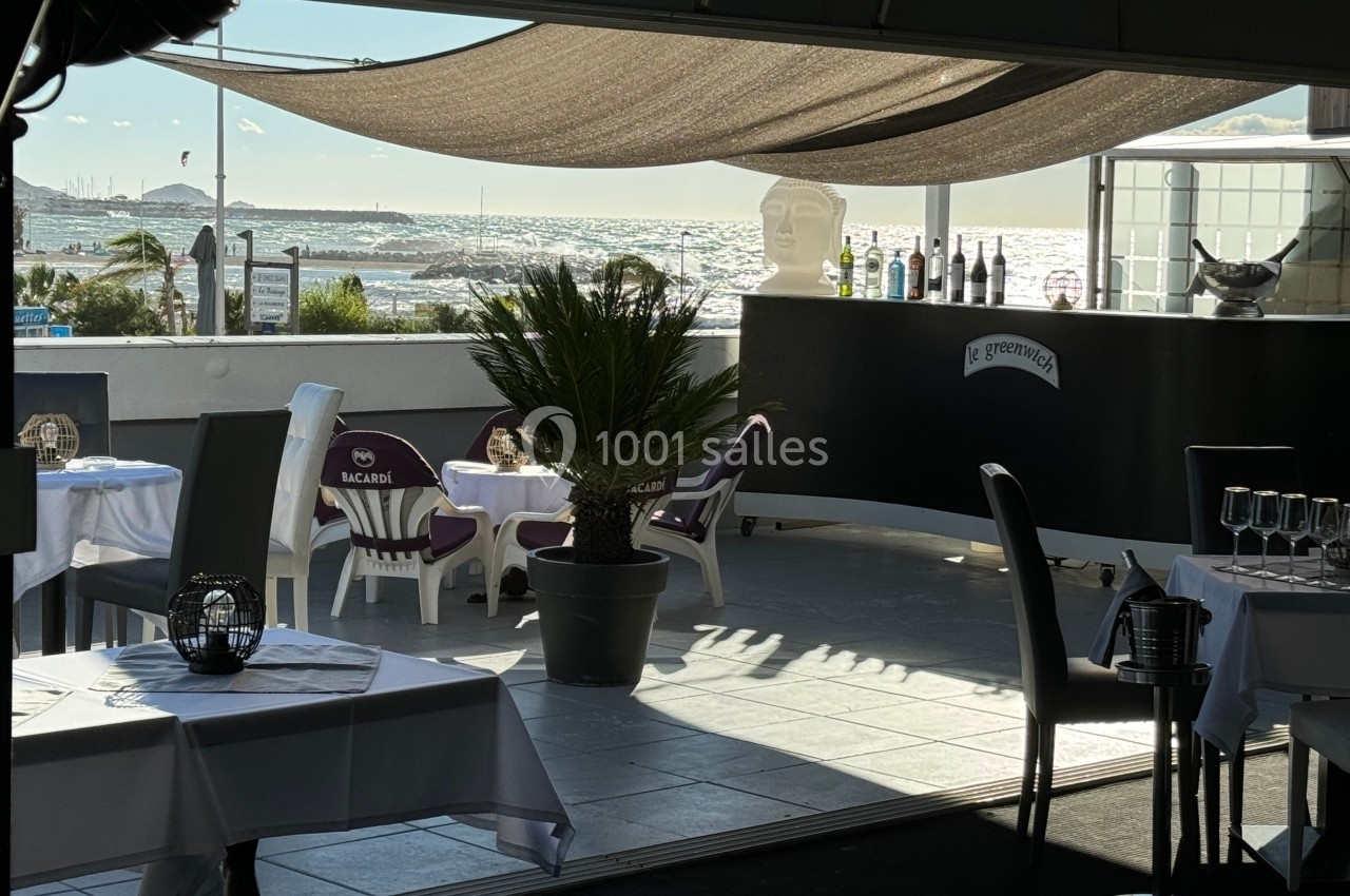 Terrasse d'un restaurant avec vue sur la mer, tables dressées, palmiers en pot et bar en arrière-plan.