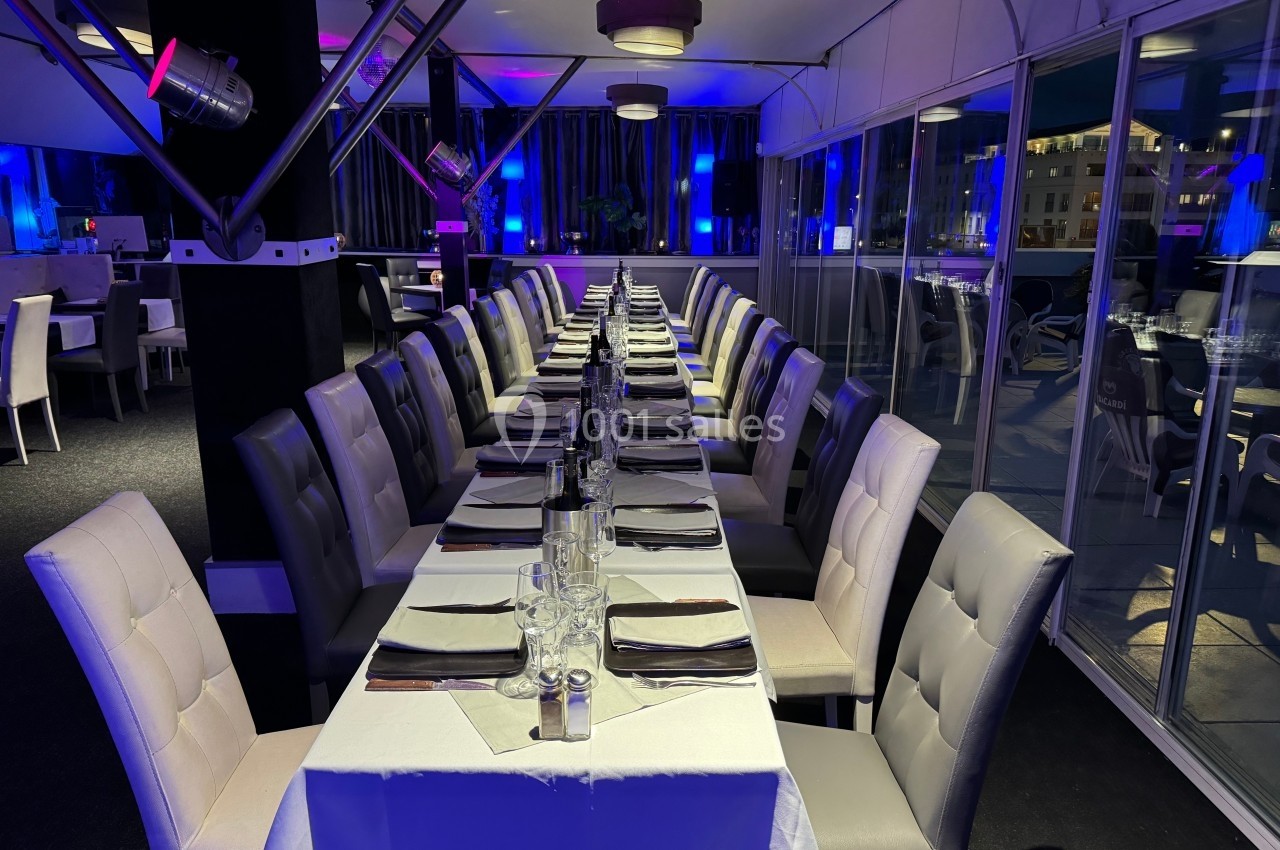 Salle de restaurant élégante avec une longue table dressée, entourée de chaises, éclairée par une lumière tamisée.