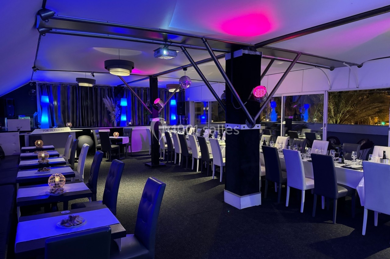 Salle de restaurant élégante avec éclairage tamisé bleu et rose, tables dressées et chaises modernes.