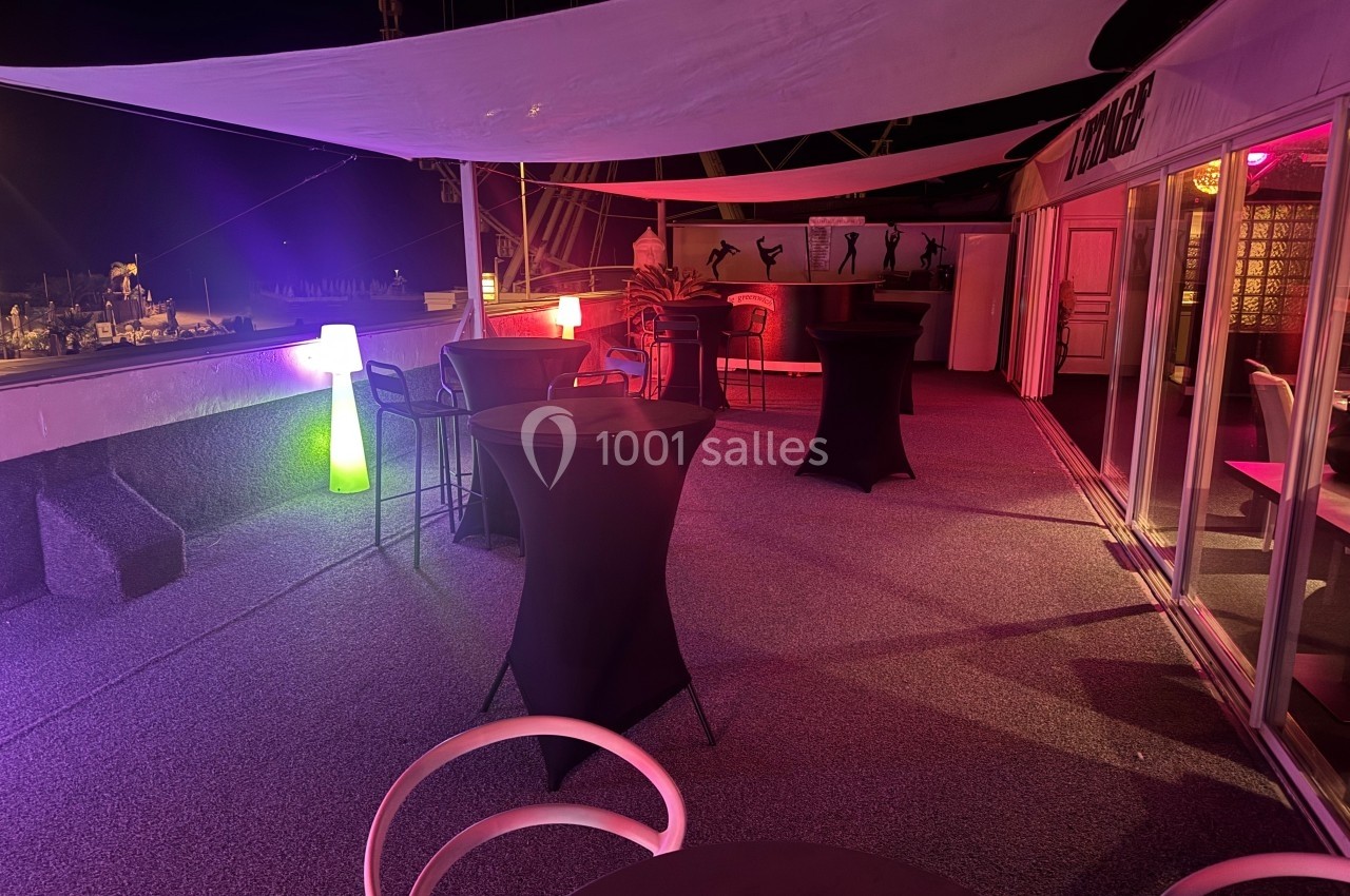 Terrasse extérieure éclairée de nuit avec tables hautes, chaises et décorations lumineuses sous des voiles tendues.