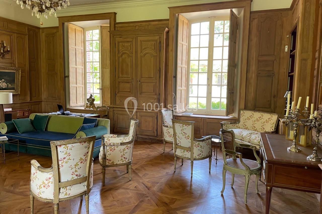 Salon élégant avec parquet en bois, fauteuils fleuris, canapé bleu, bureau ancien et grandes fenêtres lumineuses.
