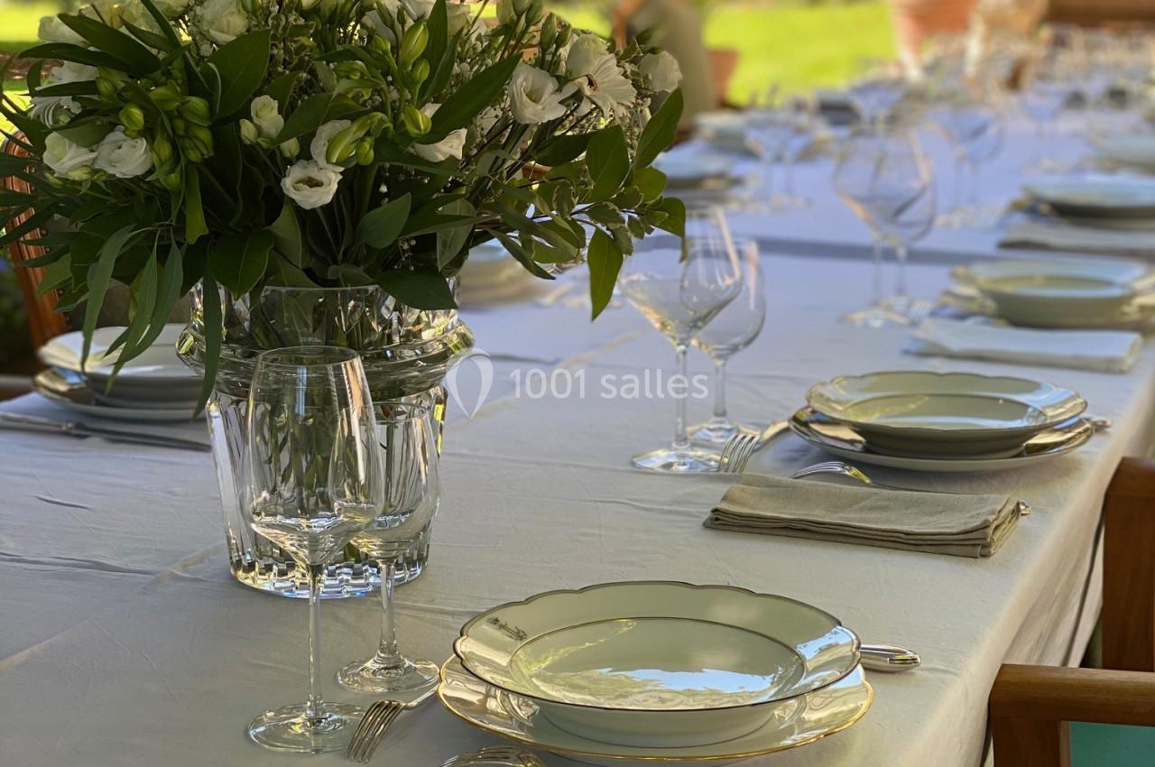 Table dressée en extérieur avec nappes blanches, vaisselle élégante, verres et bouquet de fleurs au centre.
