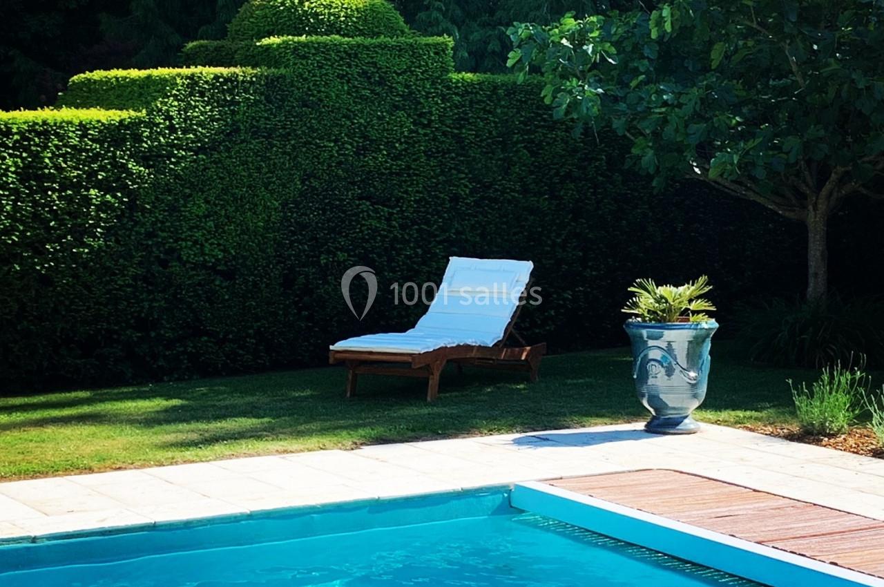 Transat blanc sur une pelouse près d'une piscine, entourée de haies taillées et d'un arbre avec un grand pot décoratif.