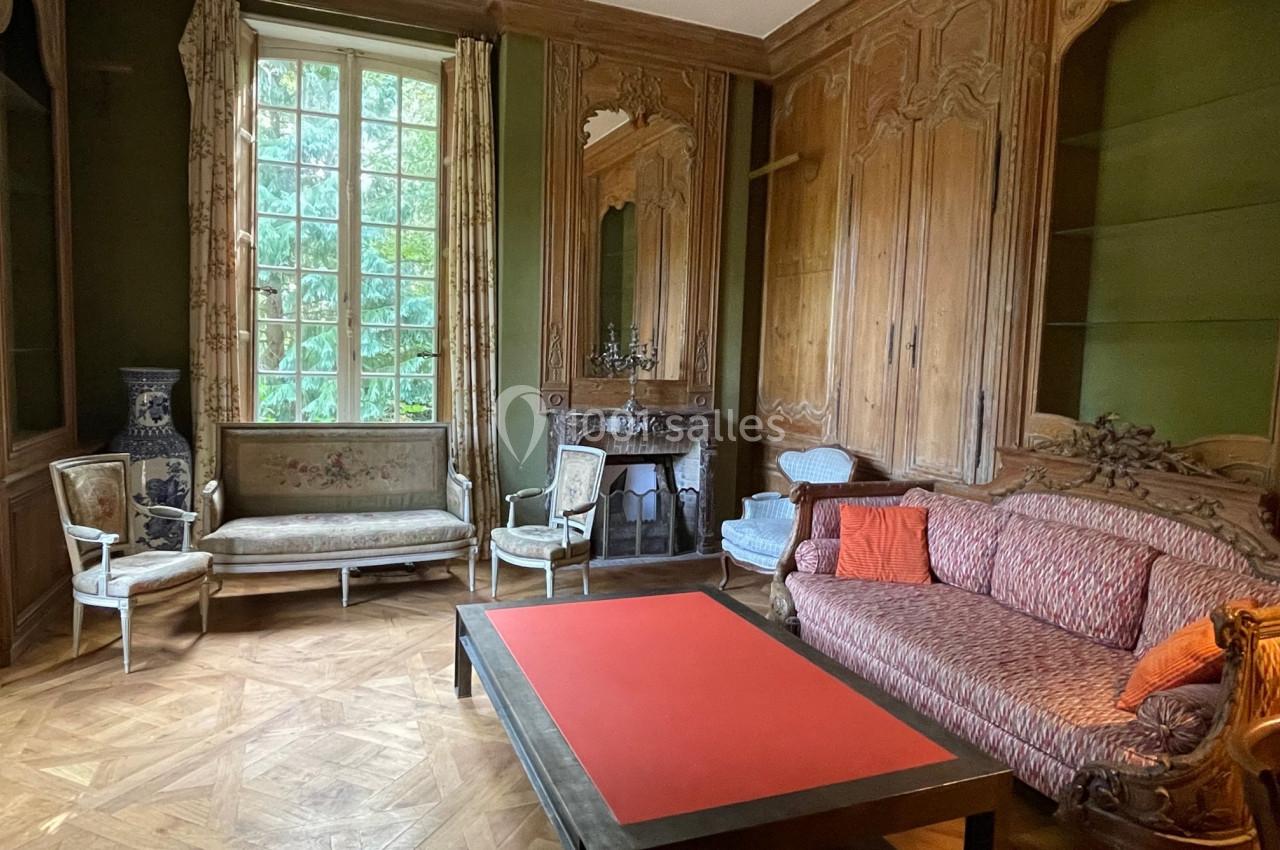 Salon ancien avec boiseries sculptées, cheminée, meubles d'époque et grande fenêtre donnant sur un jardin verdoyant.