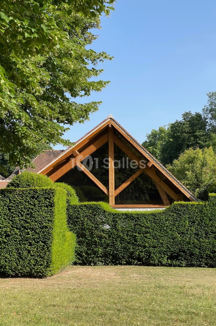 Façade en bois d'une maison avec une grande fenêtre triangulaire, entourée de haies taillées et d'arbres.