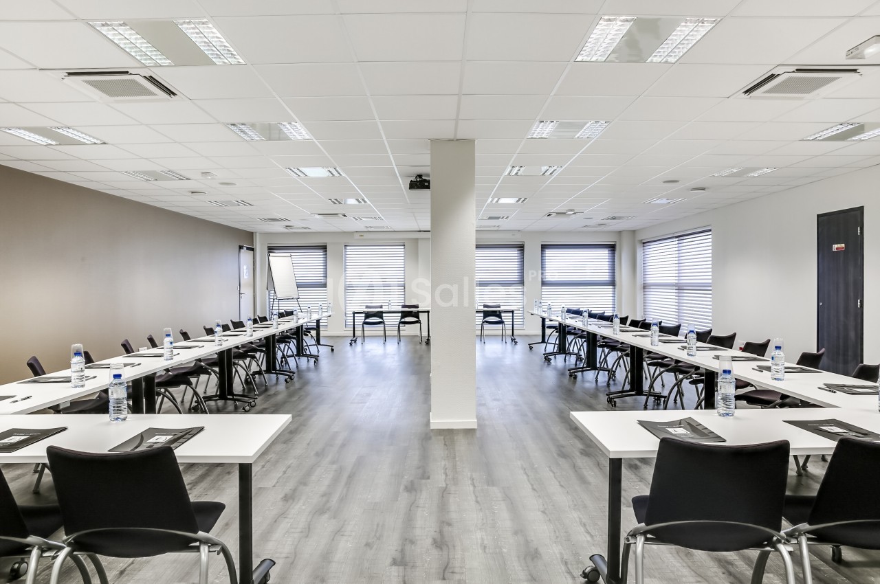 Salle de réunion lumineuse avec des tables disposées en U, chaises noires et bouteilles d'eau sur les tables.