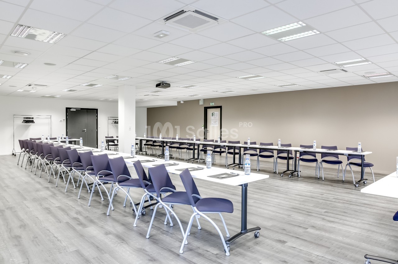 Salle de réunion lumineuse avec des tables alignées, chaises violettes et bouteilles d'eau disposées sur les tables.