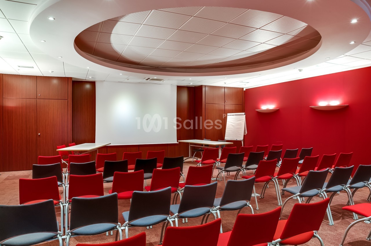 Salle de conférence avec des chaises rouges alignées, un écran de projection, un tableau blanc et des murs en bois et rouge.