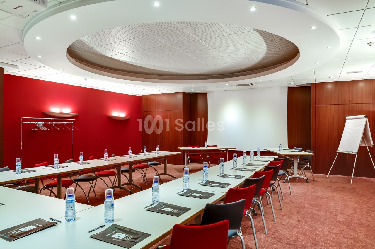Salle de réunion avec tables disposées en U, chaises rouges, tableau blanc et bouteilles d'eau sur les tables.