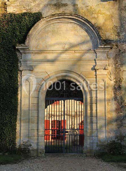 Porte en pierre voûtée avec grille en fer forgé, donnant sur une cour ensoleillée et partiellement ombragée.