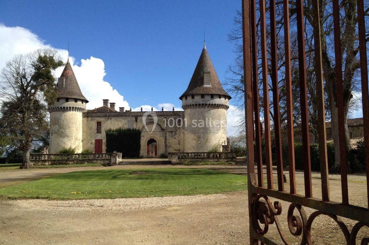 Château ancien avec deux tours rondes, vu depuis une grille en fer forgé ouverte, sous un ciel bleu.