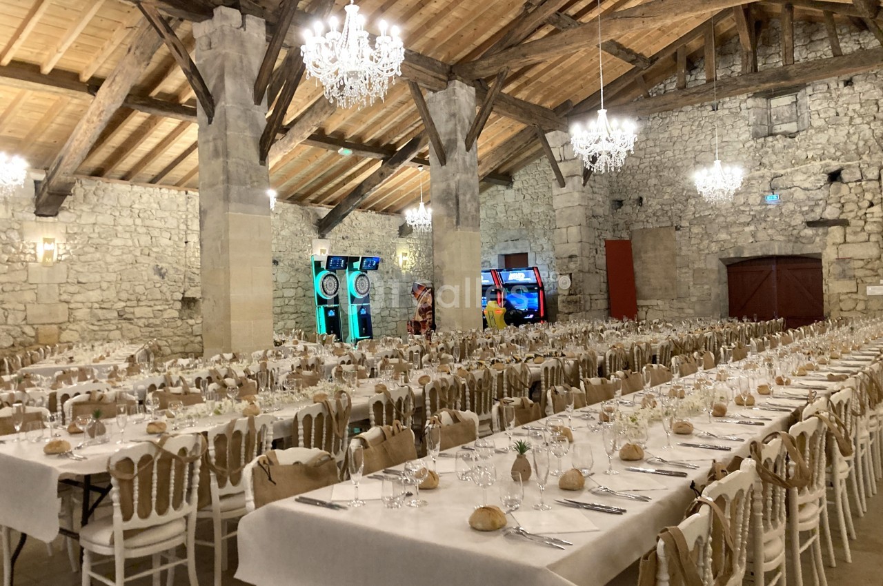 Grande salle en pierre avec tables dressées pour un repas, chaises alignées et machines de jeux en arrière-plan.