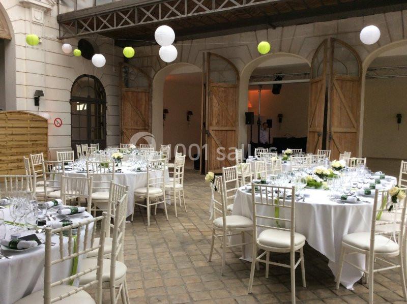 Salle de réception avec tables rondes décorées de nappes blanches, chaises en bois clair et lanternes suspendues.