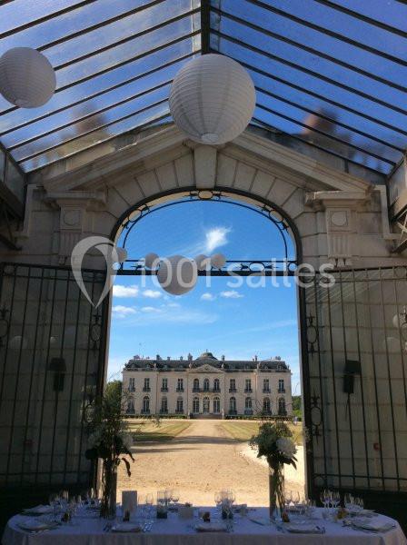 Vue d'un château à travers une verrière ornée de lanternes blanches, avec une table dressée au premier plan.