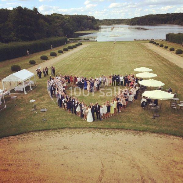 Groupe de personnes formant un cœur sur une pelouse, avec des parasols, une tente et un plan d'eau en arrière-plan.