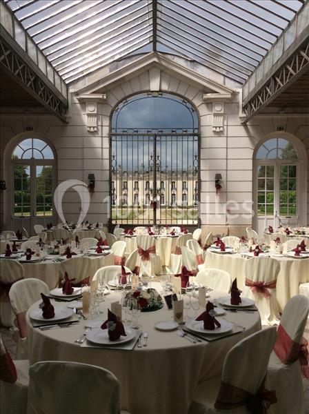 Salle de réception élégante avec tables rondes dressées, nappes blanches et décorations bordeaux, sous une verrière…