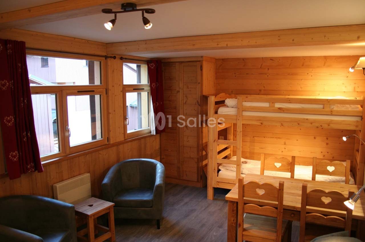 Chambre en bois avec lits superposés, fauteuils, table basse et fenêtres donnant sur l'extérieur.