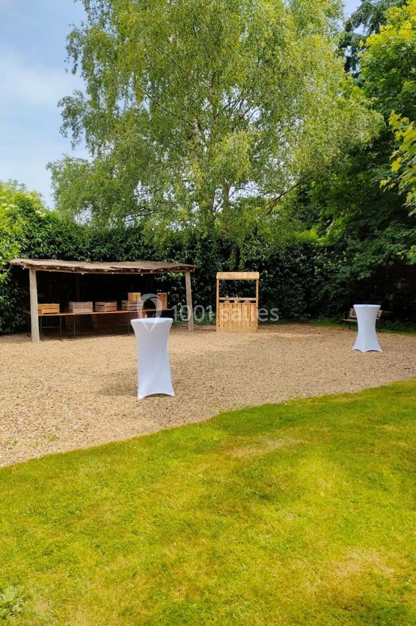 Espace extérieur avec pelouse, tables hautes drapées de blanc et structures en bois sous un arbre.