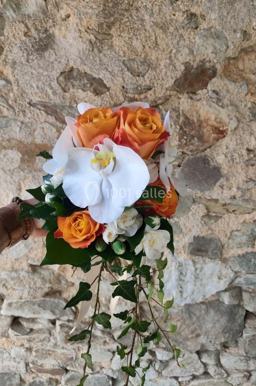 Bouquet de roses orange, orchidées blanches et feuillage, tenu devant un mur en pierre beige.