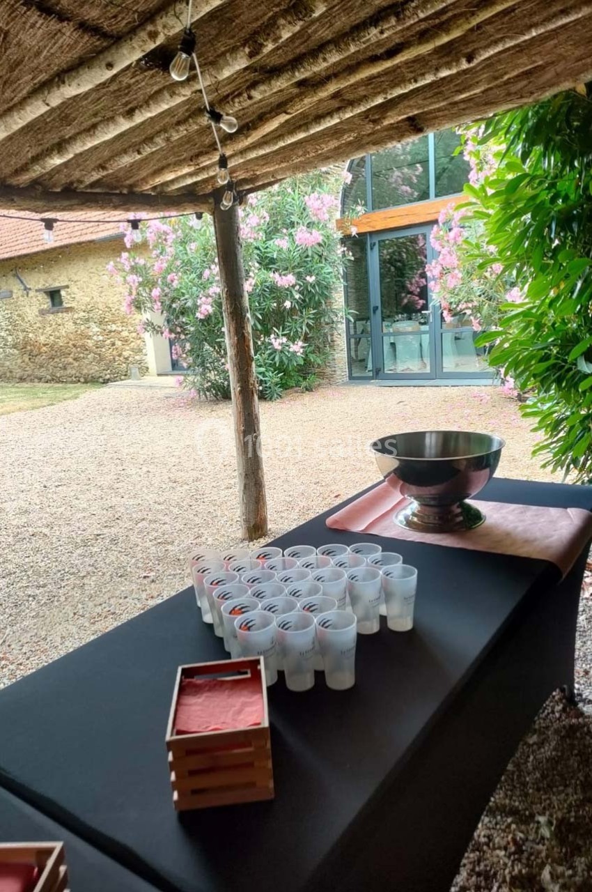 Table avec verres en plastique alignés et une grande coupe métallique, sous un abri en bois dans un jardin fleuri.