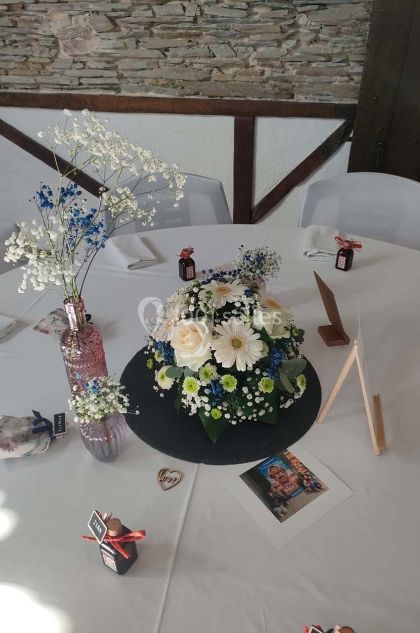 Centre de table avec bouquet de fleurs blanches et bleues, décorations et photo posés sur une nappe blanche.