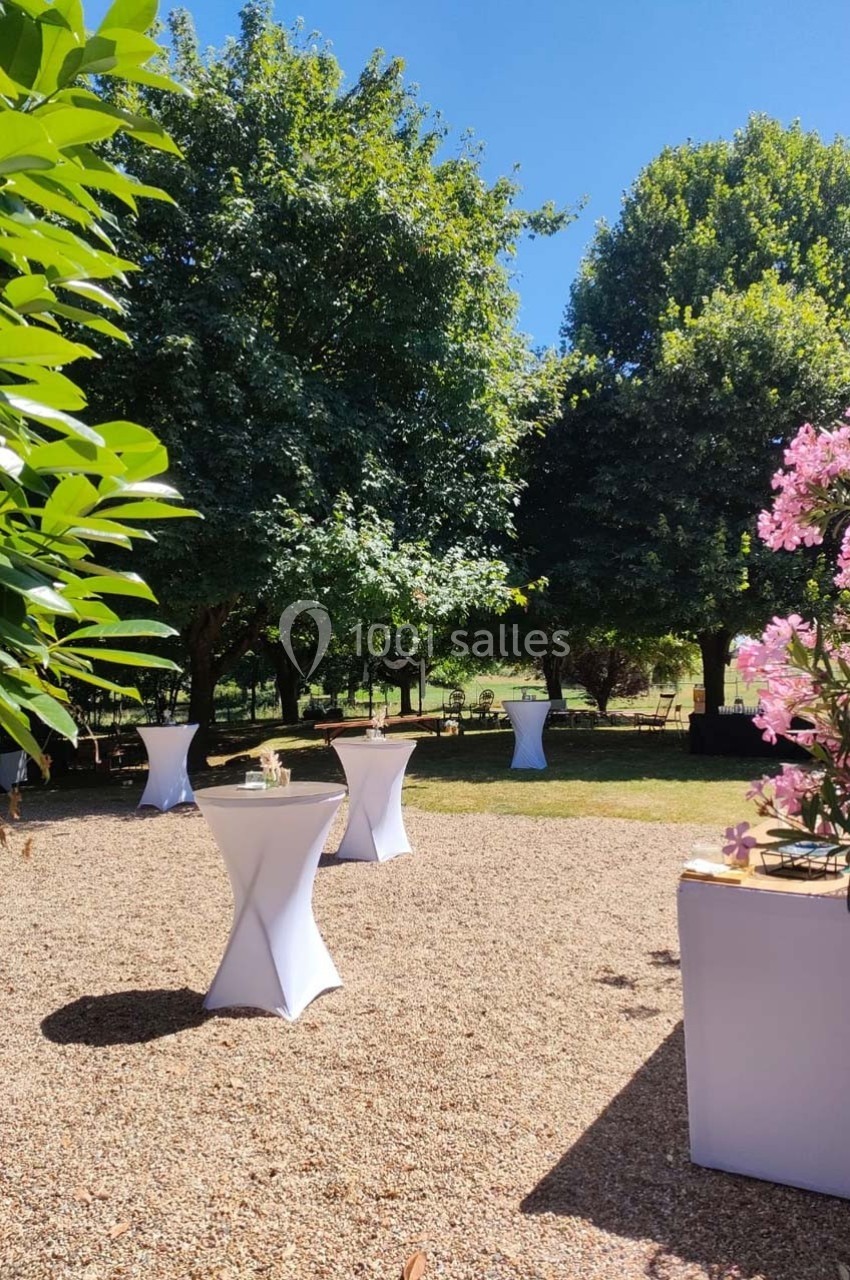 Jardin ensoleillé avec tables hautes drapées de blanc, entouré de verdure et de fleurs roses.