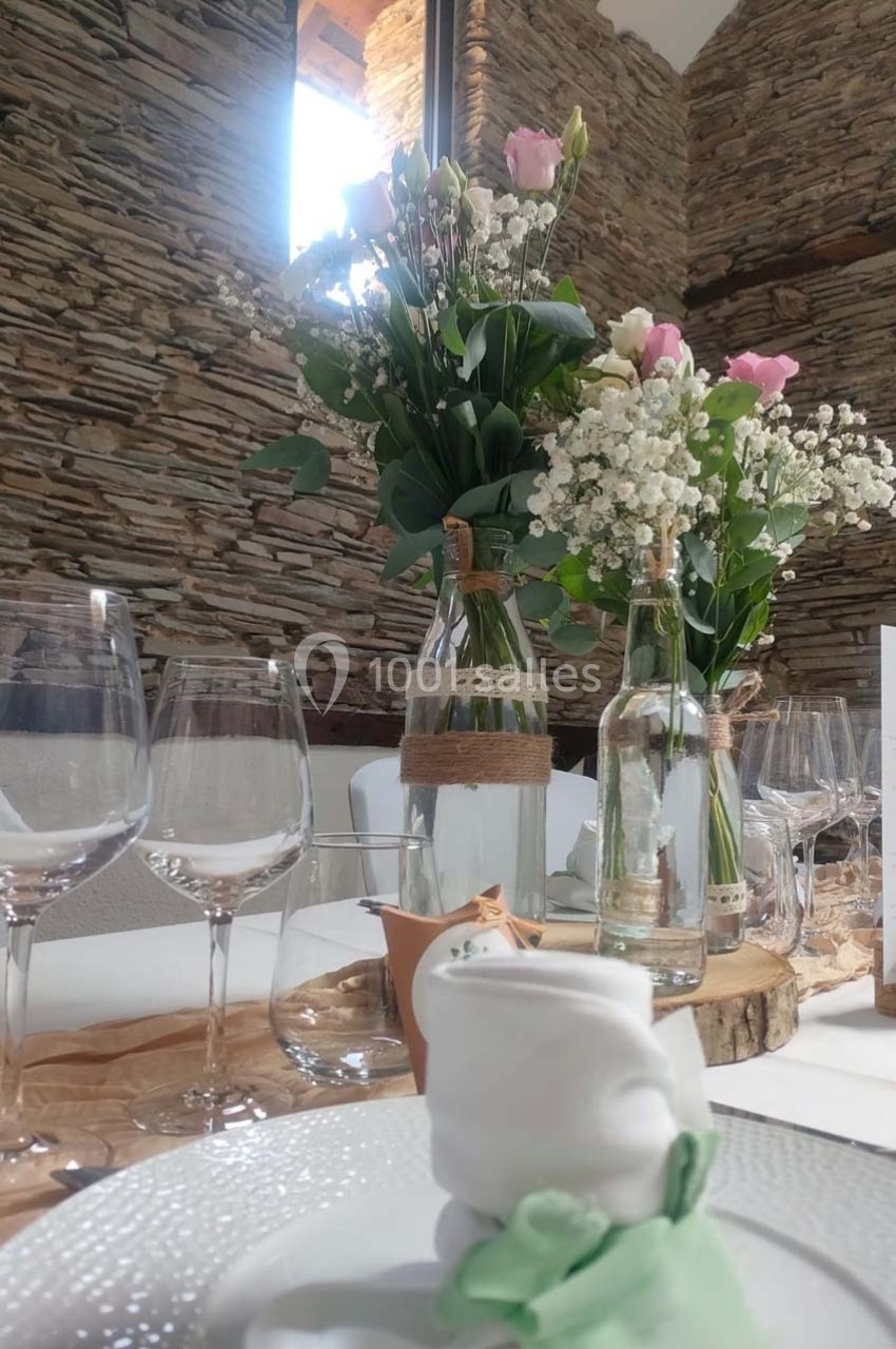 Table décorée avec des fleurs, verres et serviette pliée, dans une salle aux murs en pierre apparente.