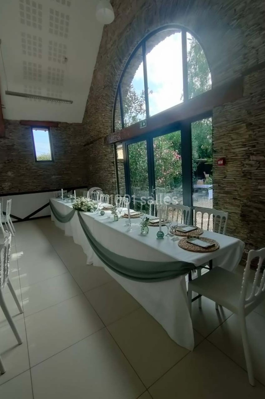 Salle de réception lumineuse avec une grande baie vitrée, table décorée de nappes blanches et guirlandes vertes.