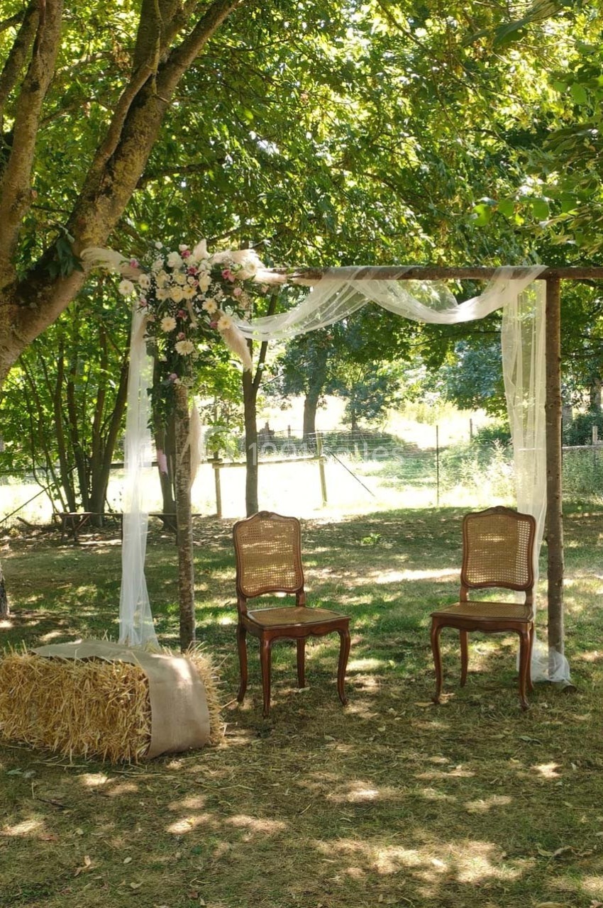 Arche en bois décorée de voilages et de fleurs, avec deux chaises en osier et une botte de paille, dans un jardin ombragé.