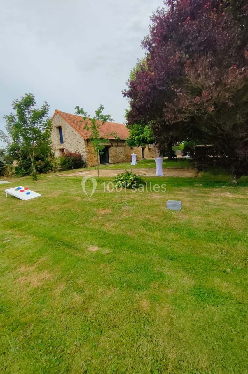 Pelouse avec un jeu de lancer, tables hautes blanches et une maison en pierre entourée d'arbres.