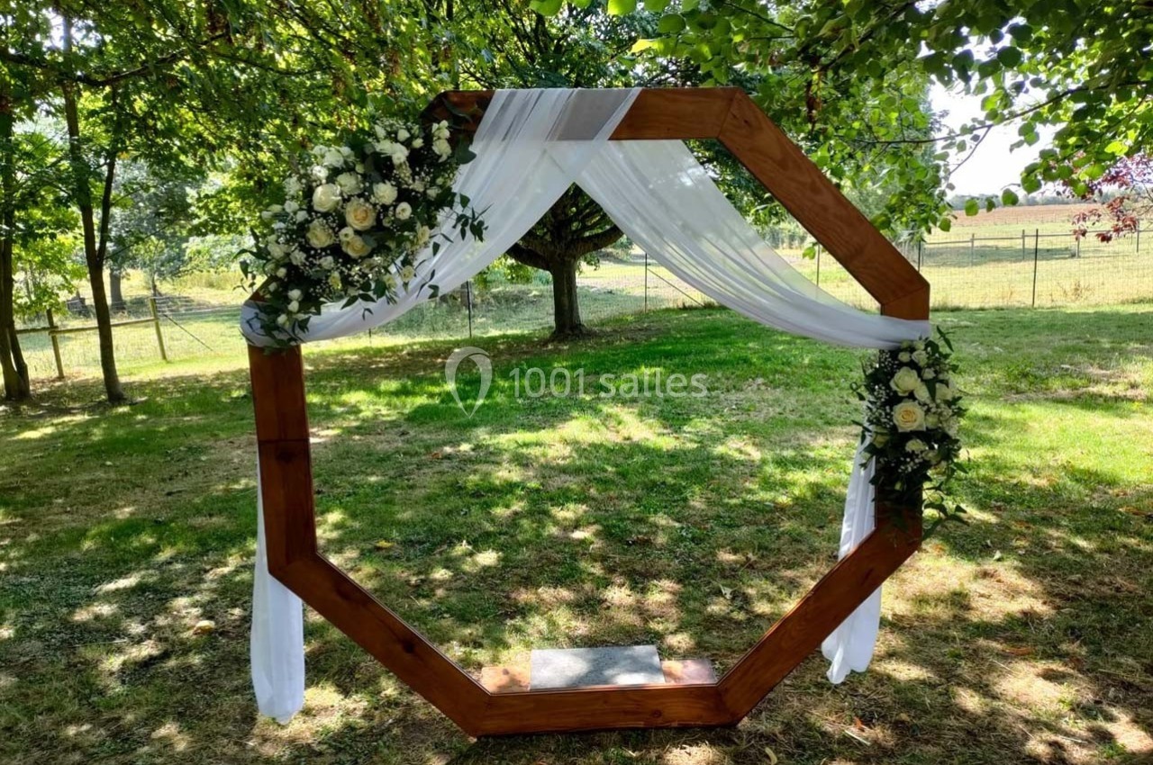 Arche octogonale en bois décorée de fleurs blanches et de voilages, installée dans un jardin verdoyant.