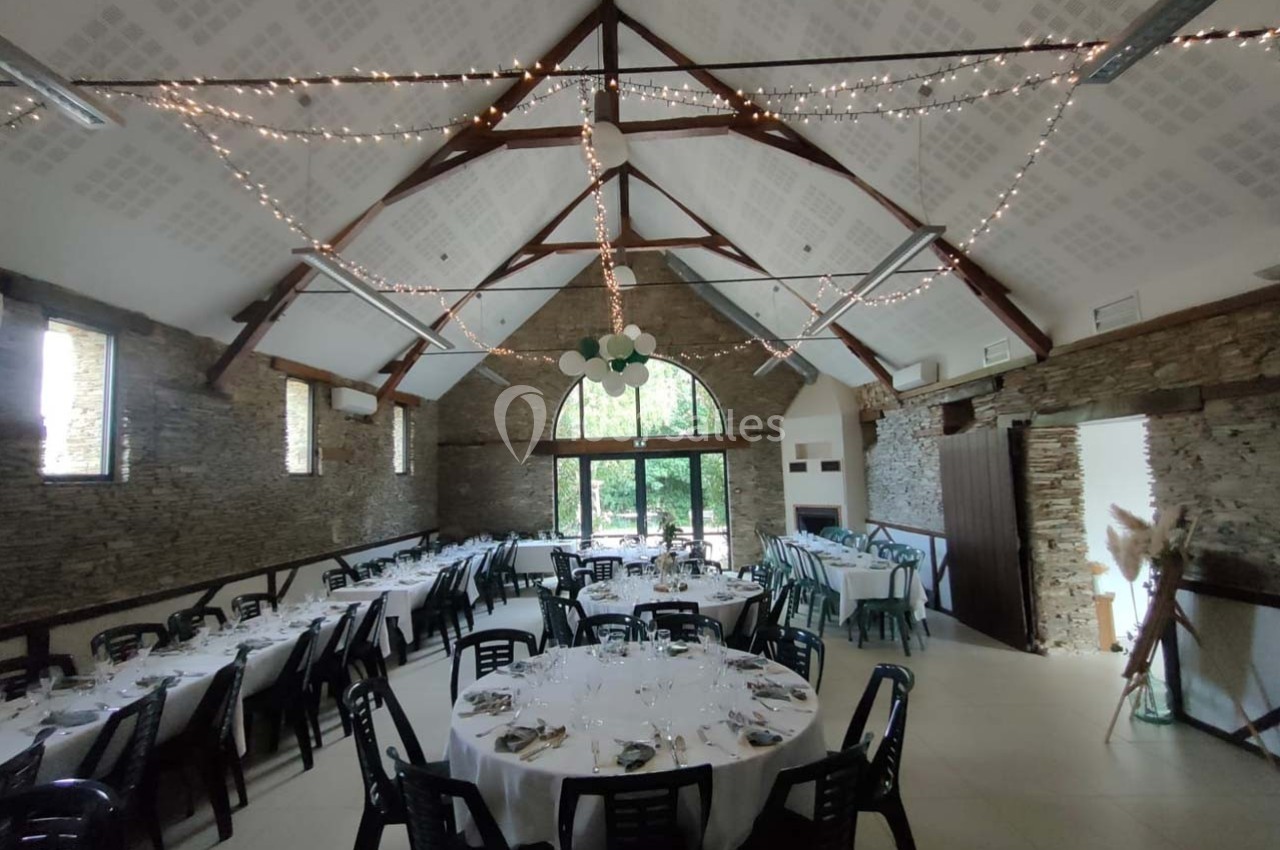 Salle de réception avec murs en pierre, tables rondes dressées et guirlandes lumineuses suspendues au plafond.