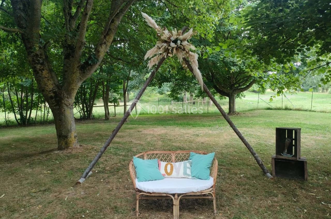 Banc en rotin décoré de coussins, placé sous une arche triangulaire ornée de fleurs séchées, dans un jardin verdoyant.