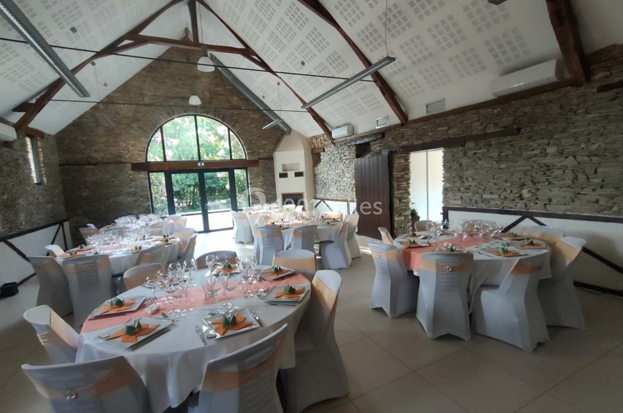 Salle de réception décorée avec des tables rondes dressées, nappes blanches et serviettes colorées, éclairée par une grande…