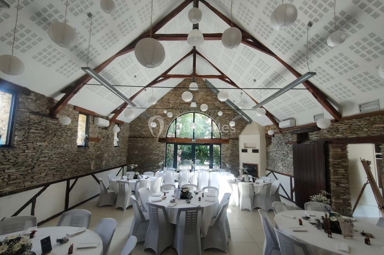 Salle de réception lumineuse avec murs en pierre, tables rondes dressées et lanternes suspendues au plafond.