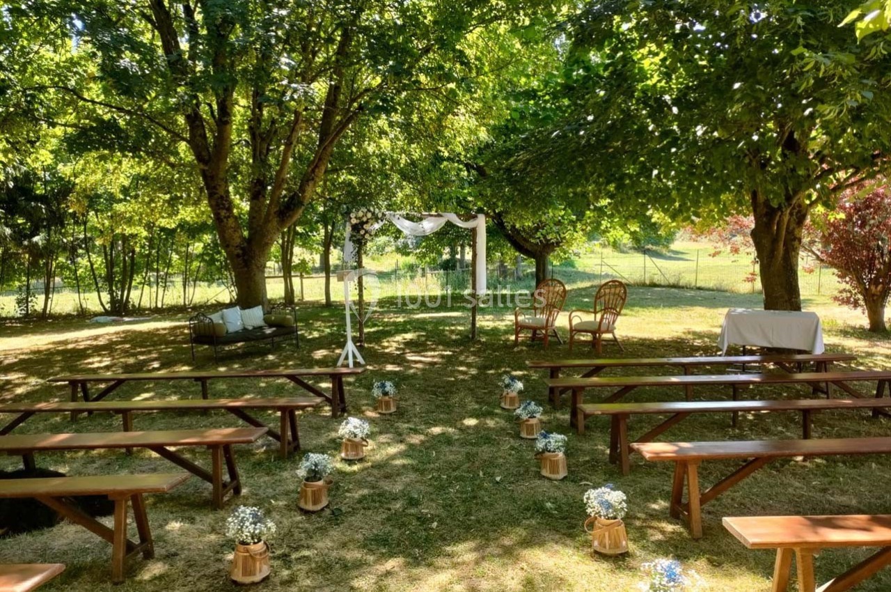 Cérémonie en plein air avec bancs en bois, arche décorée et chaises sous des arbres dans un cadre verdoyant.