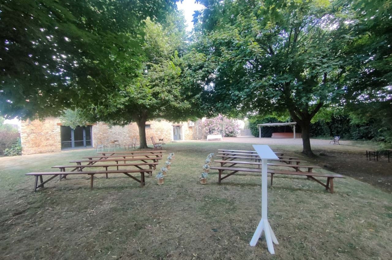 Jardin avec bancs en bois disposés pour un événement, entouré d'arbres et d'un bâtiment en pierre.