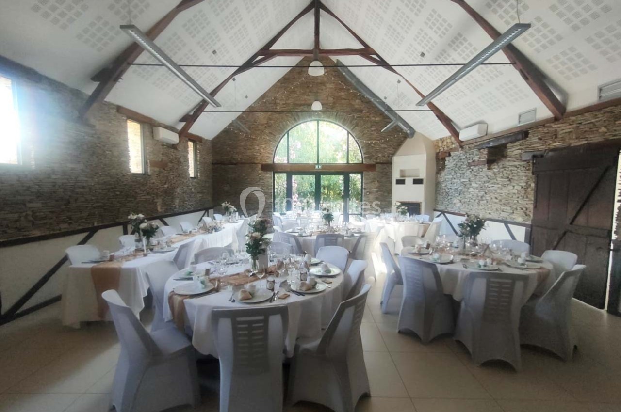 Salle de réception lumineuse avec tables rondes dressées, nappes blanches et décorations florales, murs en pierre apparente.