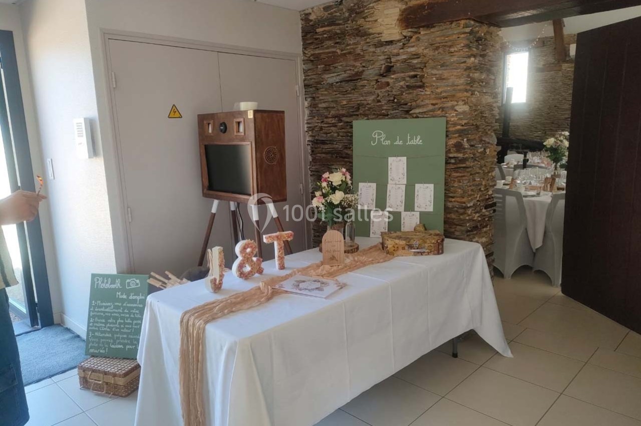 Table décorée pour un mariage avec urne, plan de table, fleurs et photobooth en bois dans une salle lumineuse.