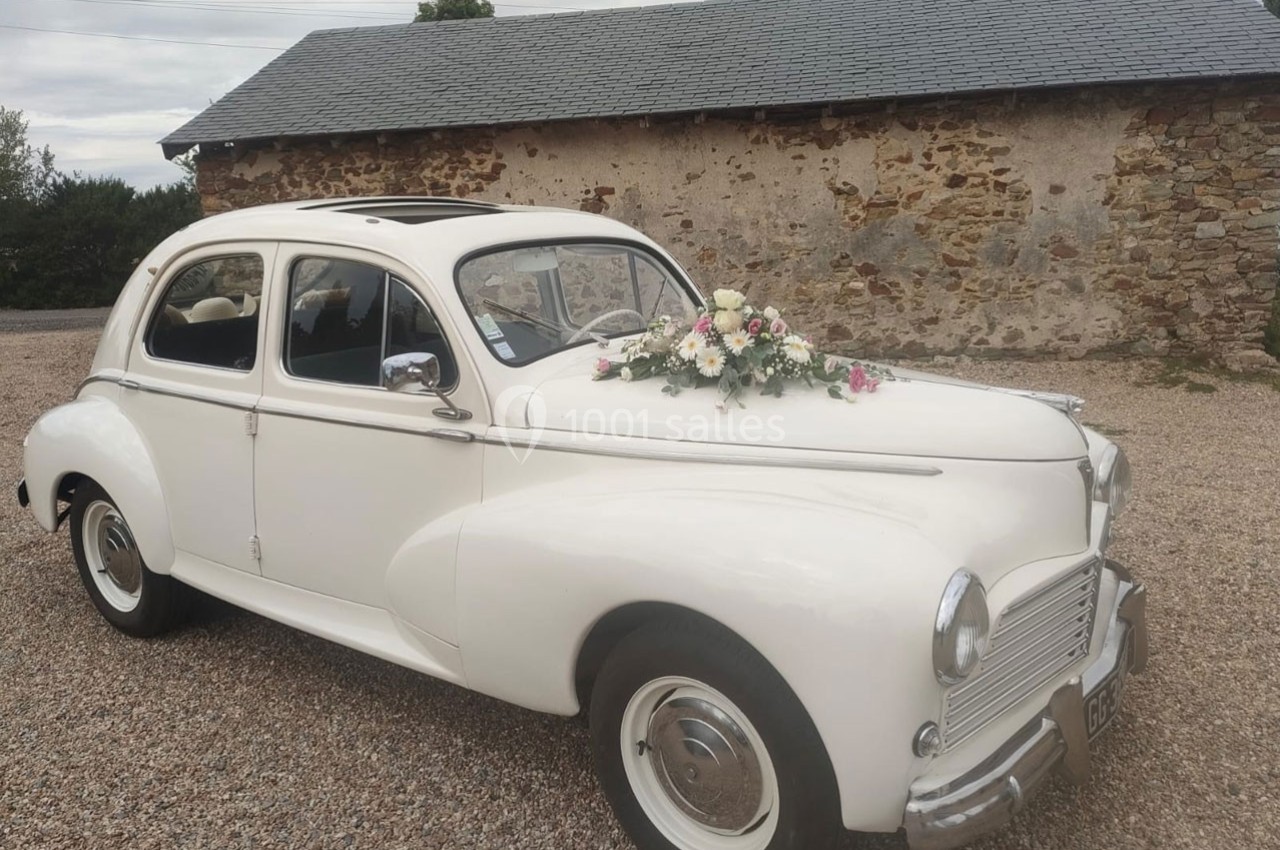 Voiture ancienne blanche décorée de fleurs, stationnée devant un bâtiment en pierre avec un toit en ardoise.